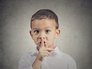 Closeup portrait young serious child, boy placing finger on lips as if to say, shhh, be quiet, silence, isolated grey wall background. Facial expression, human emotions, signs, symbols, body language